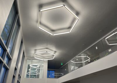 Modern building interior with large hexagonal LED ceiling lights, glass railings, white walls, and tall windows letting in natural light.