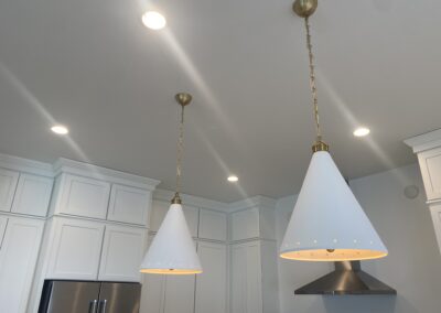 Two cone-shaped pendant lights with brass chains hang from a white kitchen ceiling, surrounded by recessed lights and white cabinetry.