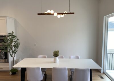 A modern dining room with a white table, six white chairs, a vase with greenery, a minimalist light fixture, and a large window to the right.