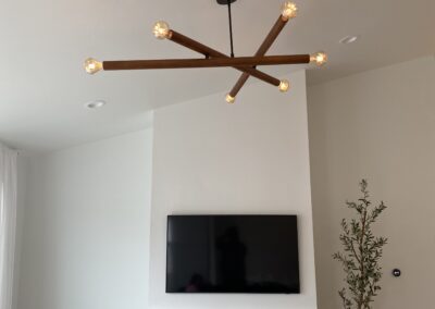 A modern living room with a wooden sputnik chandelier, a wall-mounted TV above a white fireplace, a small side table with a lamp, and a potted plant in the corner.