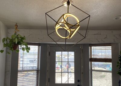 Geometric pendant light fixture with glowing rings hangs from the ceiling in a room with large windows, a door, and some indoor plants.