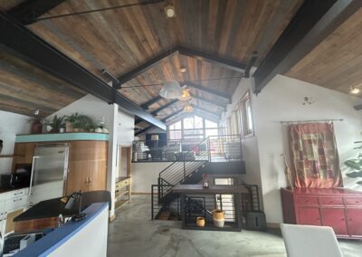 Open-plan loft with wood ceiling, modern kitchen, metal railings, large windows, and eclectic decor including plants and colorful textiles.