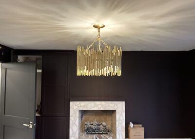 A gold chandelier hangs from the ceiling above a marble fireplace in a dark-walled room with an open door and a cardboard box on the floor.