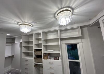 A modern walk-in closet with built-in white shelves, drawers, and two ceiling chandeliers; a window shows greenery outside.
