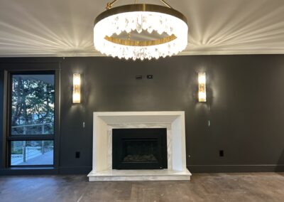 Modern living room with dark walls, a white marble fireplace, two wall sconces, and a large round chandelier. A window to the left shows outdoor greenery.