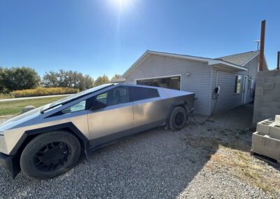 A Tesla Cybertruck is parked on a gravel driveway and charging from an electric charger mounted on the side of a house under a clear blue sky.
